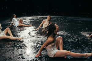 Photographie de quatre danseuses dansant au sol, assises sur la scène recouverte d'eau