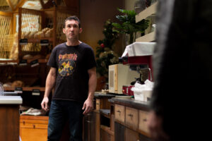 Dans une boulangerie, un homme est debout devant les baguettes de pain et fait face à un autre personnage que l'on voit de dos.