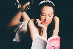Audrey Vernon portant une courrone de fleurs et en train de lire son propre livre "Comment épouser un milliardaire