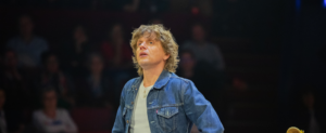 Photo portrait d'Alex Lutz sur scène. Il est vêtu d'un ensemble en jean, regarde au loin avec les mains sur la taille.