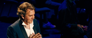 Photo portrait d'Alex Vizorek devant un pupitre et entouré d'un orchestre.