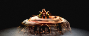 Quatre porteurs en train de faire voltiger une acrobate. Autour d’eux, d’autres artistes effectuent des mouvements circulaires créant un effet visuel de vitesse et de flou sur la photo.