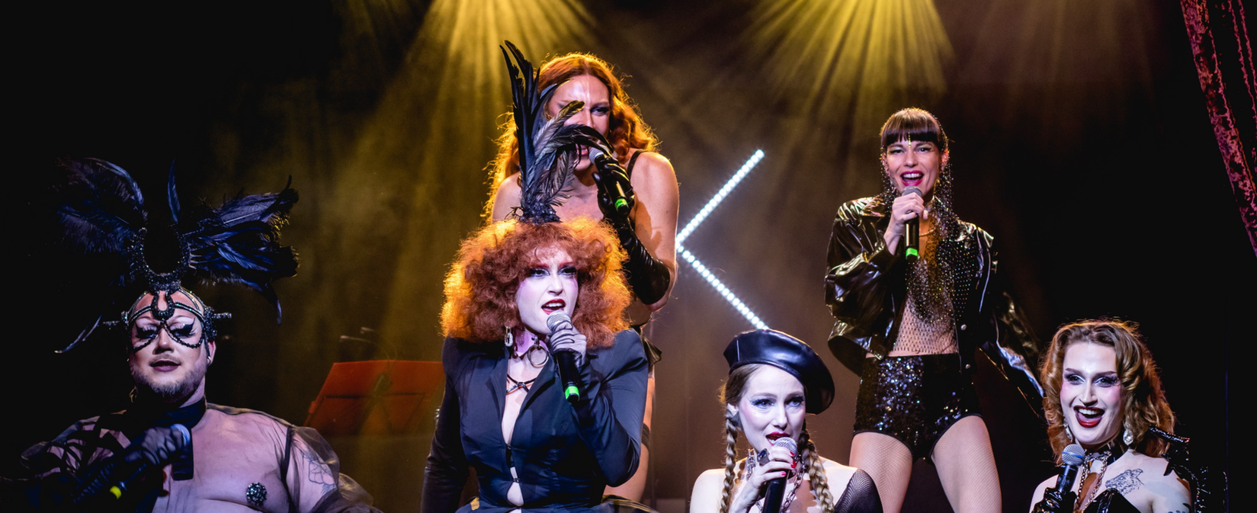 Cinq artistes sur scène en train de chanter à l'aide de micros. Leur tenue de scène est noire avec des paillettes.