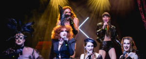 Cinq artistes sur scène en train de chanter à l'aide de micros. Leur tenue de scène est noire avec des paillettes.