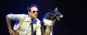 Un comédien avec des lunettes de soleil et une main sur la hanche se tient à côté d'un chien sur scène.