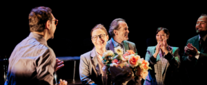 Quatre comédiens en costard cravate applaudissent et semblent féliciter une comédienne située au centre et tenant un bouquet de fleurs.