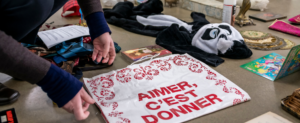 Visuel montrant plisueurs objets disposés sur le sol (une peluche panda, un jeu pour enfants, un livre...) et au centre un sac avec inscrit "aimer c'est donner" et des main qui le déposent.