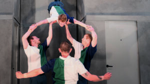 Trois acrobates en portent un troisième qui se retrouve en équilibre la tête en bas.
