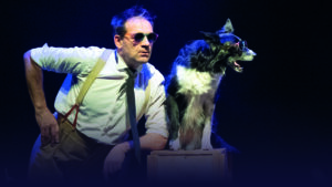 Un comédien avec des lunettes de soleil et une main sur la hanche se tient à côté d'un chien sur scène.